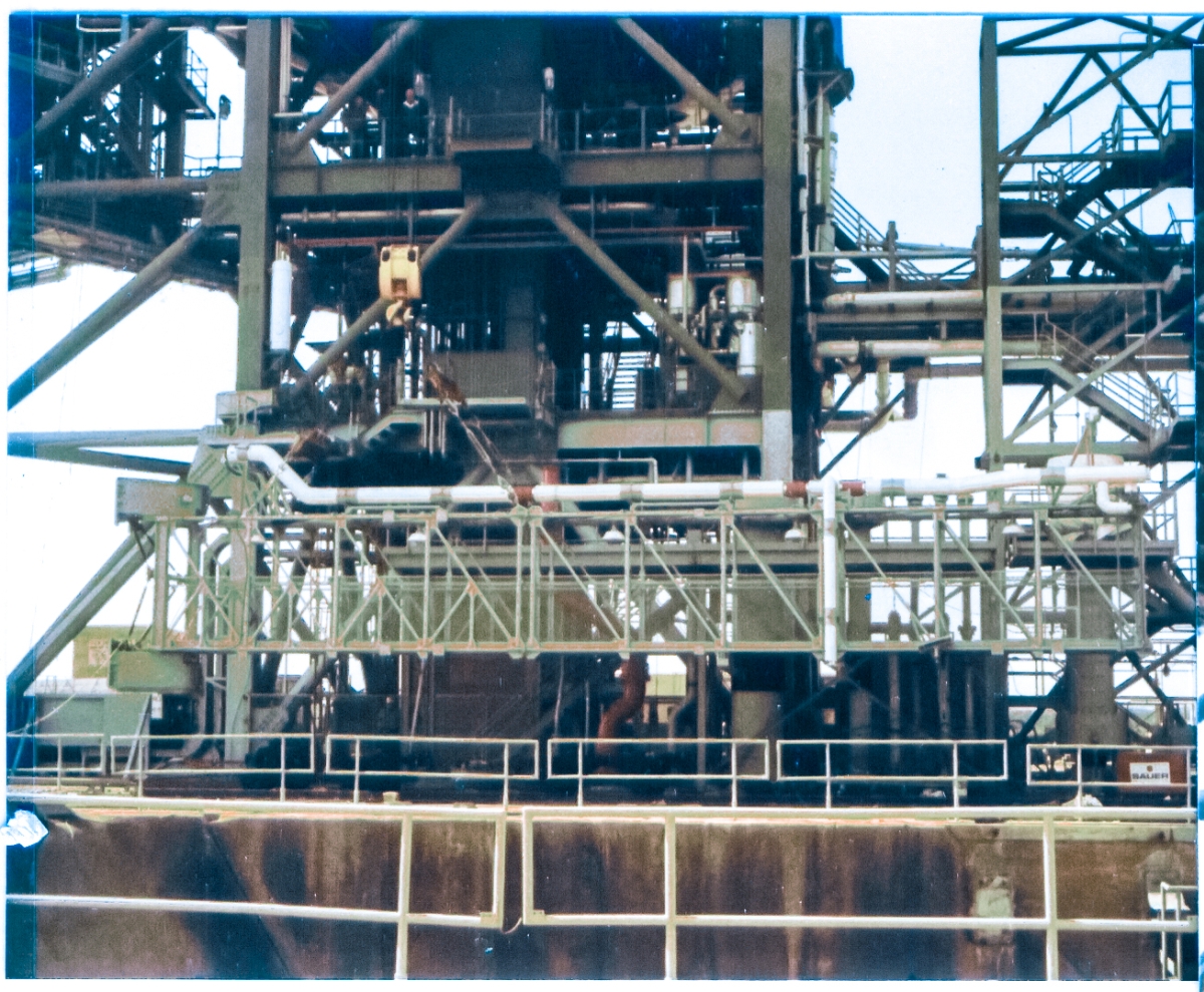 The weight of the Gox Arm Truss is no longer borne by its support stanchions, and instead is held in suspension by the Hammerhead Crane, just above the pad deck, while the ironworkers give everything a final series of inspections, before it begins its vertical journey of over twenty stories to where it will be attached to the Fixed Service Structure at Space Shuttle Launch Complex 39-B, Kennedy Space Center, Florida.